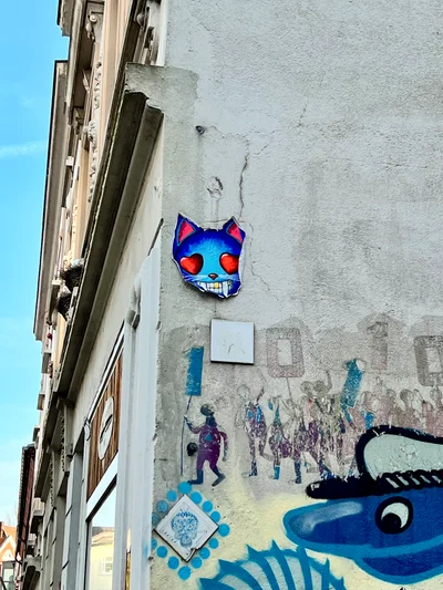 Ein blaues Katzengesicht mit Herzaugen als Street-Art an einer Hauswand mit weiteren Wandmalereien