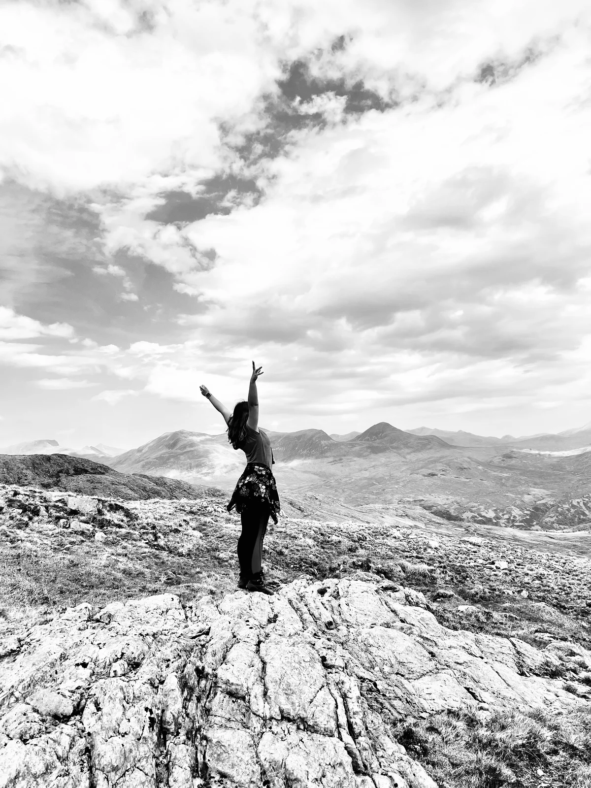 Eine Person steht mit erhobenen Armen auf einem Berggipfel, Schwarz-Weiß-Aufnahme vor weitem Bergpanorama