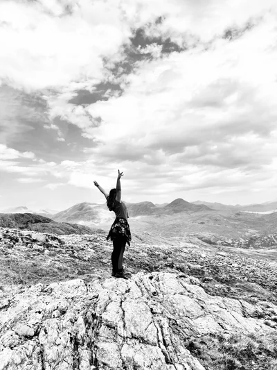 Eine Person steht mit erhobenen Armen auf einem Berggipfel, Schwarz-Weiß-Aufnahme vor weitem Bergpanorama