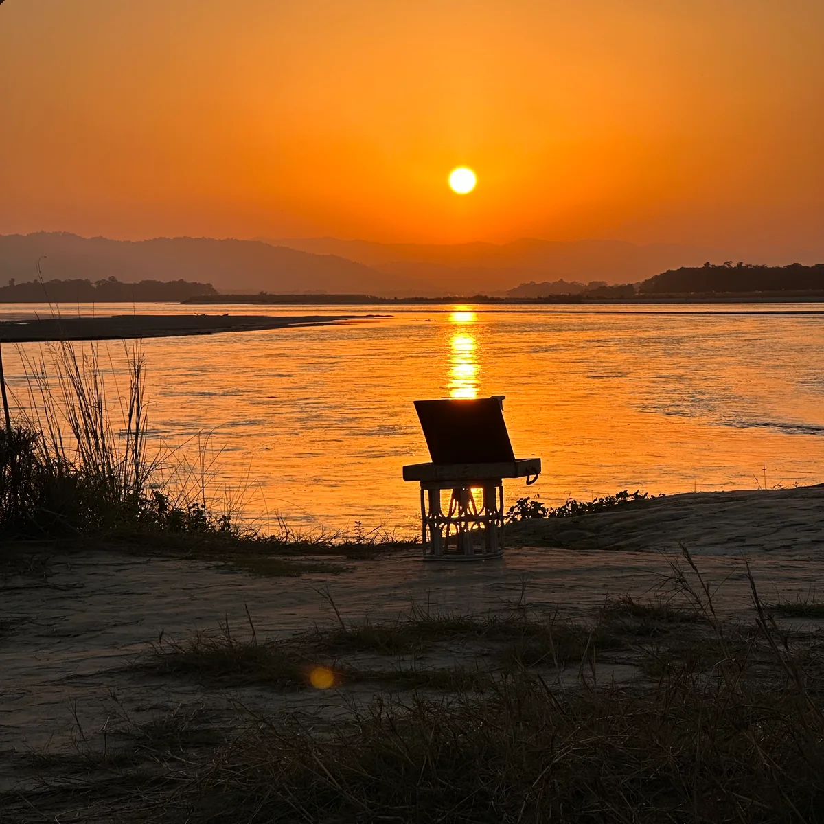 Ein goldener Sonnenuntergang spiegelt sich auf einem breiten Fluss, ein einzelner Stuhl am Ufer