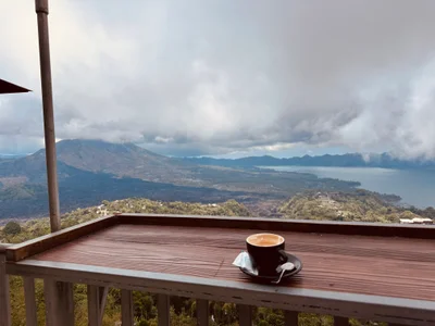 Eine Kaffeetasse auf einem Holzgeländer mit Blick auf einen Vulkan und See unter Wolken
